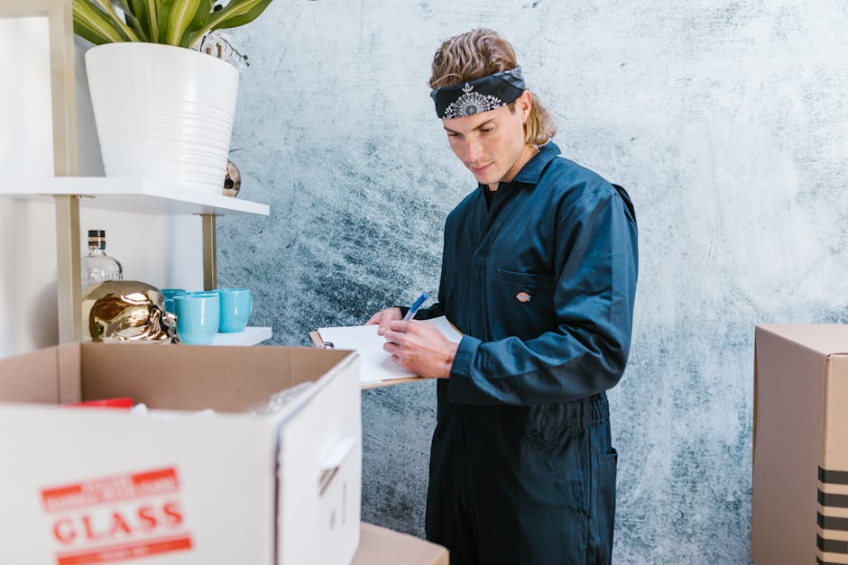 A young man with light skin and shoulder-length blond hair, wearing a black bandana around his forehead and a navy blue work jumpsuit, is standing indoors in front of a textured light blue wall. He is holding a clipboard and pen, appearing to take inventory or note details related to house removals or packing in preparation for a home relocation. To his left, there are cardboard boxes, one labeled with 'CLASS' in red, and partially visible packing materials inside. Behind him, on white shelving, there are decorative items including a large white ceramic pot with a plant, blue ceramic mugs, and a small glass decanter. The scene is well-lit with natural or artificial light, emphasizing the professional environment of a removal company's storage or packing area, supporting the context of furniture transport and moving logistics associated with Alexandra Palace landmark removals.