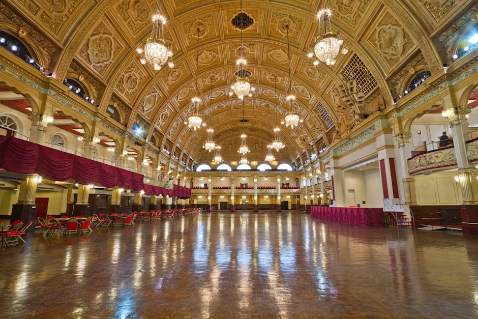 Interior view of a large, ornate hall with an elaborately decorated vaulted ceiling featuring intricate moldings and multiple large chandeliers hanging from above. The space has polished wooden flooring reflecting the lighting, with red upholstered chairs arranged along one side, and maroon draped curtains on the upper balcony level. To the right, a white and gold staircase leads to the balcony, with a small area cordoned off by red velvet ropes. The hall appears prepared for an event or gathering, with some packaging materials and furniture not present in this image. The environment suggests a landmark location suitable for house removals or moving services, such as temporary storage or packing at Alexandra Palace, with no visible furniture or boxes currently being moved, but indicating the scale and interior setting for furniture transport and logistical planning. Removal Company Haringey’s services may involve handling large items in or around such venues during home relocations or large-item transfers. The image is well-lit, emphasizing the grand architectural features and spaciousness of the hall.