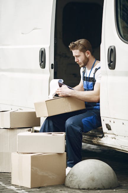 A male removal worker with short brown hair and a beard, wearing a striped t-shirt and blue overalls, is seated on the threshold of a white moving van with the sliding door open. He is holding and exa