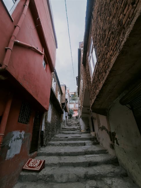 An outdoor narrow staircase composed of uneven concrete steps ascending through a densely built residential area, with closely spaced brick and plastered houses on either side. To the left, a red-painted house with a small window and a black metal grille, while the right side features a building with exposed brickwork and another window. A red foam cushion or packaging material rests at the base of the stairs near the bottom step. The area has minimal natural lighting, with the sky visible above, indicating an upward view through the tight, urban alleyway. This setting illustrates the challenging access for home relocation or furniture transport in narrow streets, where skilled removals services by companies like Removal Company Haringey are often required for efficient packing, loading, and moving goods through confined spaces such as this, aligning with house removals tips for tight streets and stairs in Crouch End N8.