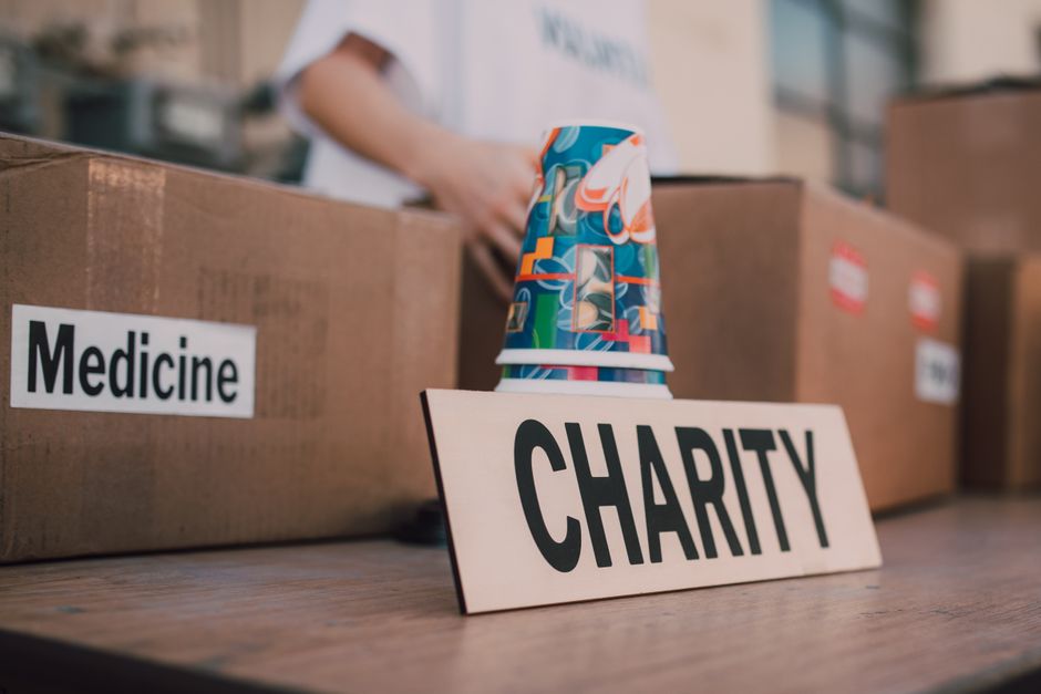 Inside a property during a home relocation, several cardboard boxes are visible on a wooden floor, with one labeled 'Medicine' and others unlabeled. In the foreground, a white sign with bold black letters reading 'CHARITY' is placed next to a colorful, patterned paper cup. A person wearing a white shirt stands behind the boxes, partially visible, handling some items as part of the packing and moving process. The scene is well-lit, indicating natural or ample indoor lighting, and suggests an organized packing or donation activity coordinated by Removal Company Haringey as part of a house removal or charity donation task, with boxes stored in an indoor space ready for transport.