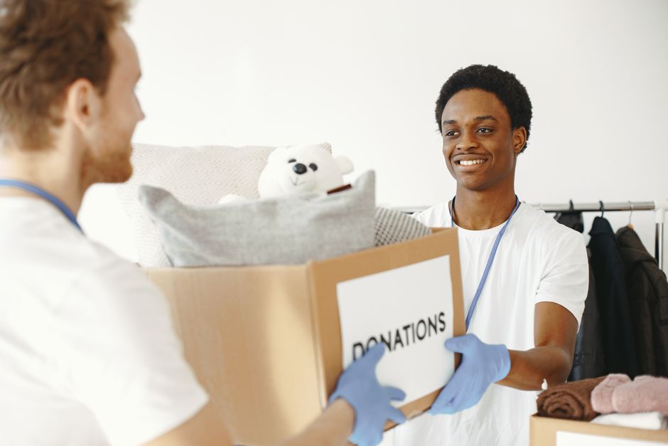 A volunteer and a man inside a home, both wearing white T-shirts and blue disposable gloves, are involved in a charity donation process related to house removals or packing and moving services. The volunteer, facing away from the camera with partial profile visible, is handing a cardboard box labeled 'DONATIONS' to the man, who is smiling and extending his hands to receive it. The cardboard box contains various items including a plush teddy bear, a folded blanket or towel, and some clothing, indicating the collection of household goods for donation. They are standing in a room with natural lighting, and in the background, there is a clothing rack with hanging garments and a light-colored sofa or cushion, suggesting a domestic environment. The image is relevant to the themes of furniture transport, packing, and efficient moving logistics typical of house removal services offered by Removal Company Haringey, supporting the website page about Muswell Hill N10 removals and charity donation tips for Haringey.