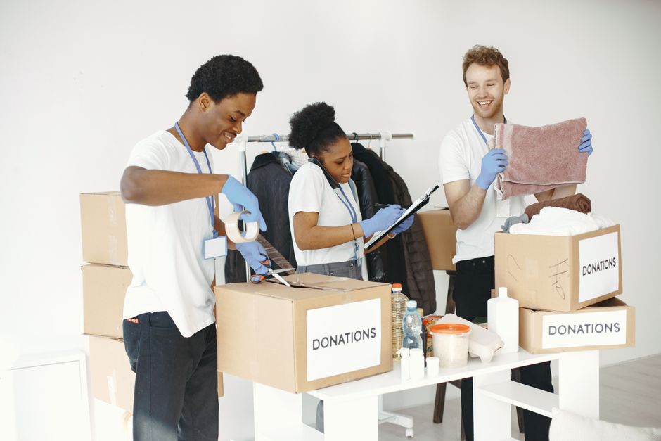 Inside a property during a home relocation, several cardboard boxes are visible on a wooden floor, with one labeled 'Medicine' and others unlabeled. In the foreground, a white sign with bold black letters reading 'CHARITY' is placed next to a colorful, patterned paper cup. A person wearing a white shirt stands behind the boxes, partially visible, handling some items as part of the packing and moving process. The scene is well-lit, indicating natural or ample indoor lighting, and suggests an organized packing or donation activity coordinated by Removal Company Haringey as part of a house removal or charity donation task, with boxes stored in an indoor space ready for transport.