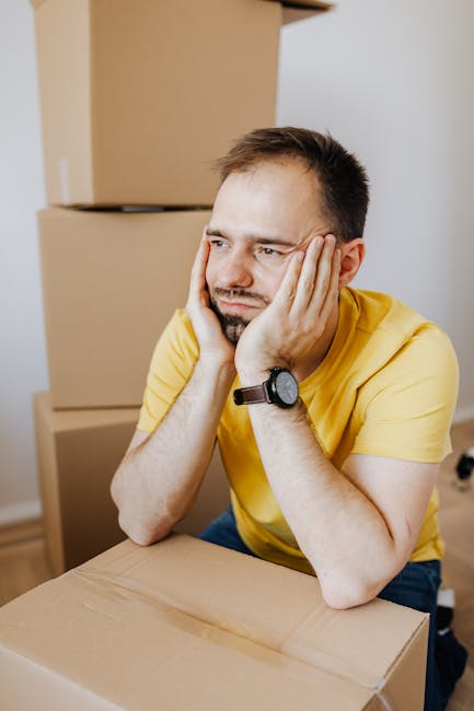 A man wearing a yellow t-shirt and a wristwatch is sitting at a table with his elbows resting on its surface, holding his face with both hands and appearing concerned. In the background, there are sev
