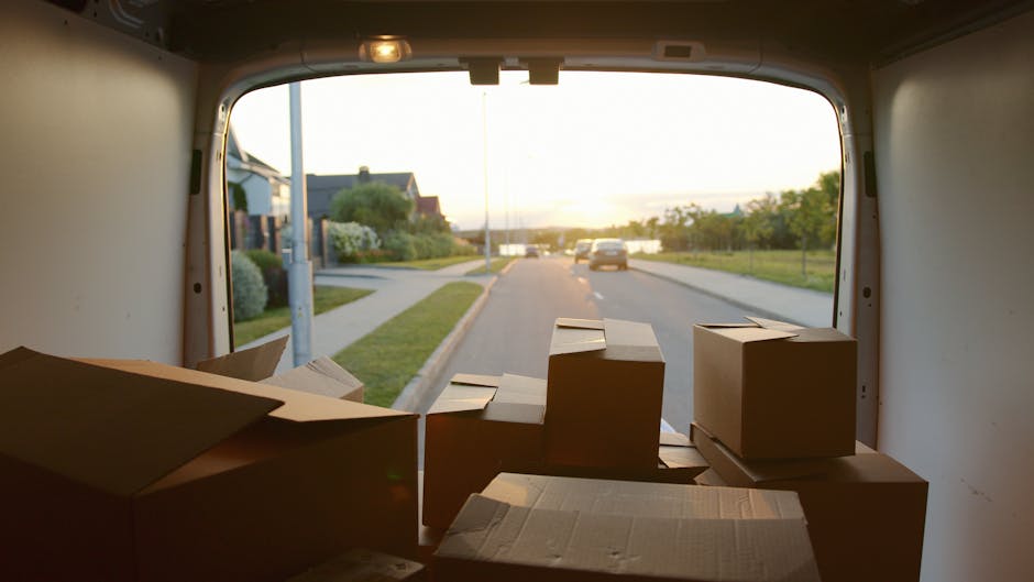 Inside the back of a van during a house removal process, with several cardboard boxes of various sizes stacked on the floor near the rear opening. The boxes are sealed with packing tape, some are partially open, and a few are placed on top of each other. The van is parked on a residential street with a sidewalk, grass verge, and houses visible in the background. The front of the van shows the exterior environment, including a parked car and trees, with the sky indicating early morning or late afternoon sunlight. This scene illustrates the loading process involved in home relocation services offered by Removal Company Haringey, highlighting the packing and transport stages of a typical move, with focus on secure packing materials and orderly loading within a moving vehicle.