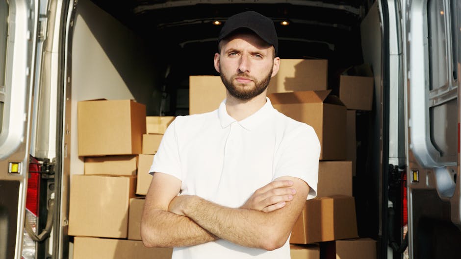A young man with a serious expression, wearing a black cap and a white polo shirt, stands inside the rear compartment of a moving van filled with stacked cardboard boxes. The boxes vary in size and are arranged closely together, some visibly wrapped in plastic or packing materials. The interior of the van is dimly lit, with the open rear doors revealing a paved area outside, possibly on High Road during a house removal or furniture transport process as part of a home relocation project. The man is positioned in front of the boxes, arms crossed, indicating involvement in packing or loading tasks supported by Removal Company Haringey, which offers professional removals and moving services in Tottenham N17.