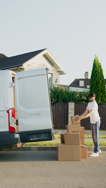 Inside the back of a van during a house removal process, with several cardboard boxes of various sizes stacked on the floor near the rear opening. The boxes are sealed with packing tape, some are partially open, and a few are placed on top of each other. The van is parked on a residential street with a sidewalk, grass verge, and houses visible in the background. The front of the van shows the exterior environment, including a parked car and trees, with the sky indicating early morning or late afternoon sunlight. This scene illustrates the loading process involved in home relocation services offered by Removal Company Haringey, highlighting the packing and transport stages of a typical move, with focus on secure packing materials and orderly loading within a moving vehicle.