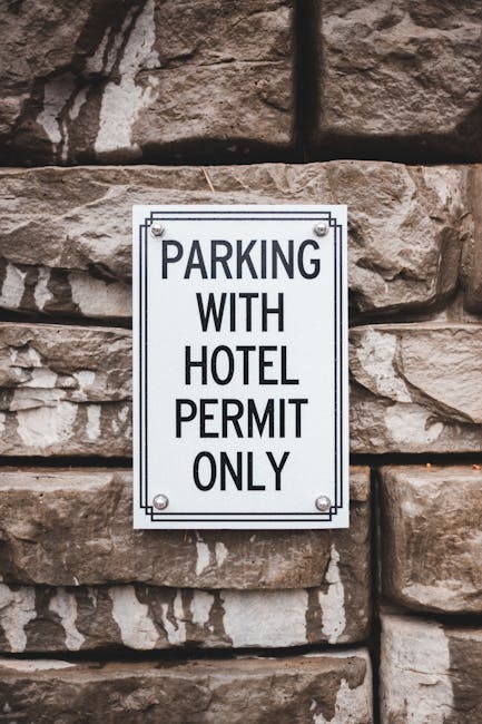 An outdoor scene showing a rectangular metal sign mounted on a rough stone wall, indicating 'PARKING WITH HOTEL PERMIT ONLY' in bold black letters. The sign is secured with four screws, one at each corner. The stone wall consists of large, irregularly shaped brownish stones with visible mortar joints. The sign is positioned at about eye level, suggesting it is meant for vehicles or pedestrians. This image is relevant to house removals and relocation services provided by Removal Company Haringey, particularly regarding parking regulations for home relocation or furniture transport in the Wood Green N22 area. The setting implies an urban environment where proper permits are essential for moving logistics, aligning with the process of planning a residential move, including vehicle access and parking permissions.