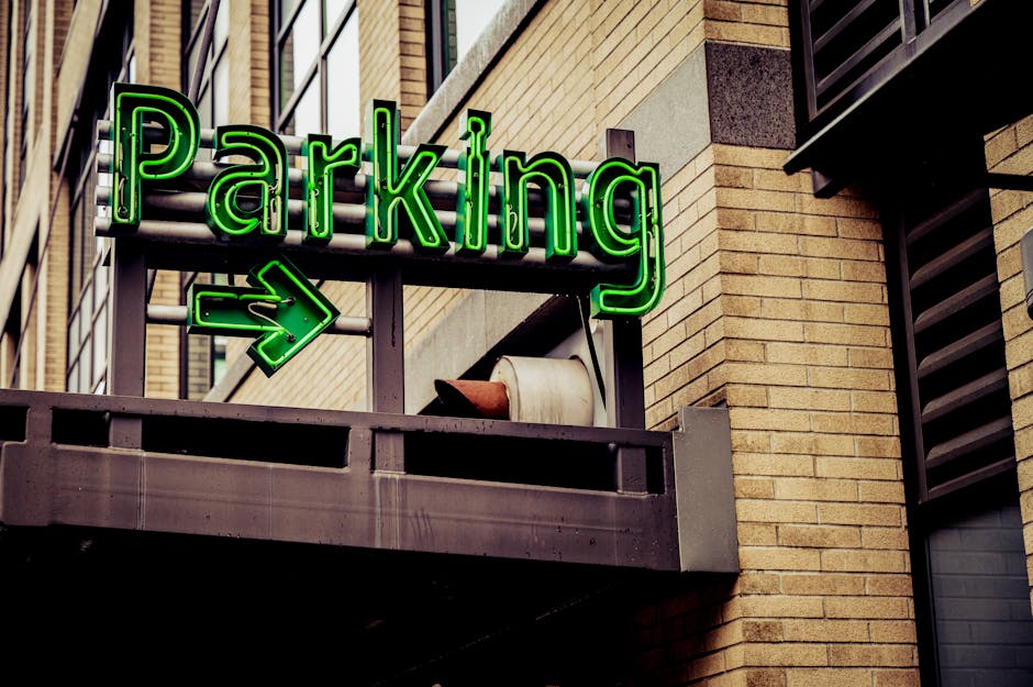 A brightly illuminated green neon sign spelling 'Parking' with a right-pointing arrow is mounted on a metal railing of an outdoor balcony. The balcony is part of a modern brick building, with yellow-brown bricks and dark window frames visible. A large, white ceramic flowerpot is placed on the railing next to the sign. Below, a cardboard box wrapped in bubble wrap, along with a plastic container and some packing materials, are visible on the balcony floor, indicating active packing or loading activity. The image is taken during daylight, with natural light illuminating the scene, which may be part of the home relocation process involving furniture transport or moving logistics, as handled by Removal Company Haringey. The overall setting reflects a typical urban residential environment, supporting indoor and outdoor moving operations.